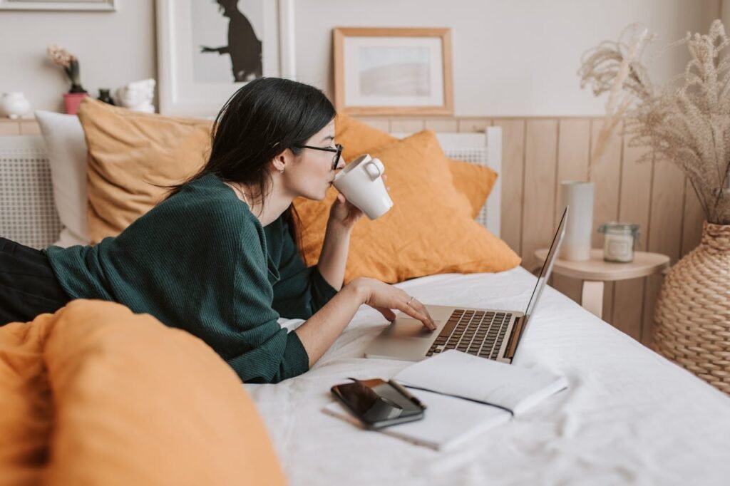 Woman relaxing on bed with coffee browsing laptop for freelance work or online learning.